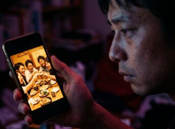 夜、暗い部屋で、昔の仲間が乾杯している写真をスマホで見つめる男性