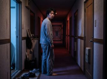 夜、自宅の廊下で振り返る部屋着の男性。廊下は同じドアが並んだまま奥へ果てしなく続いている